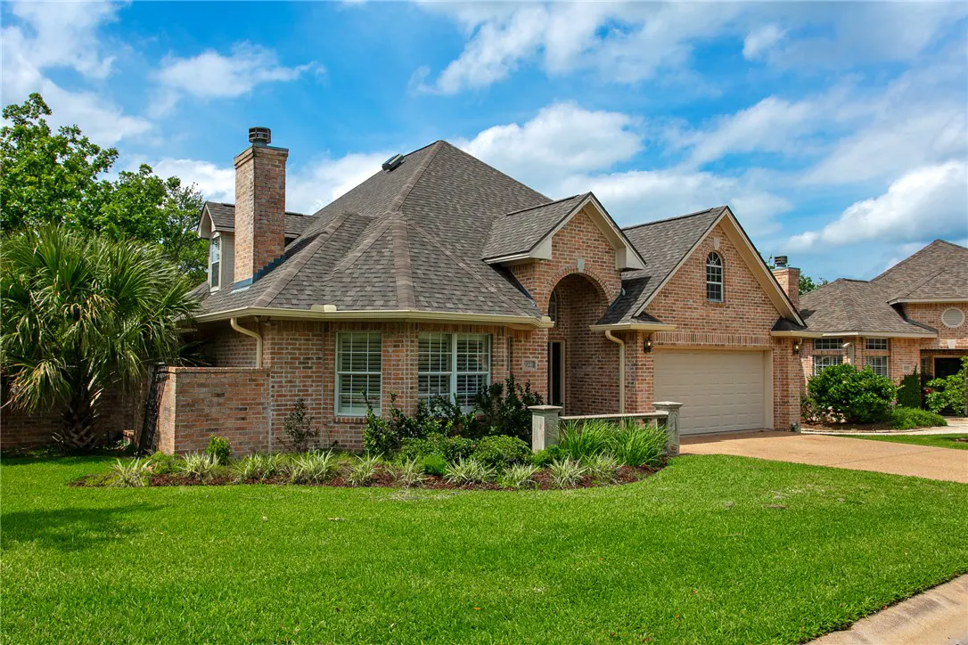 Brick house with a gray roof, chimney, and attached garage. Green lawn and blue sky with clouds.