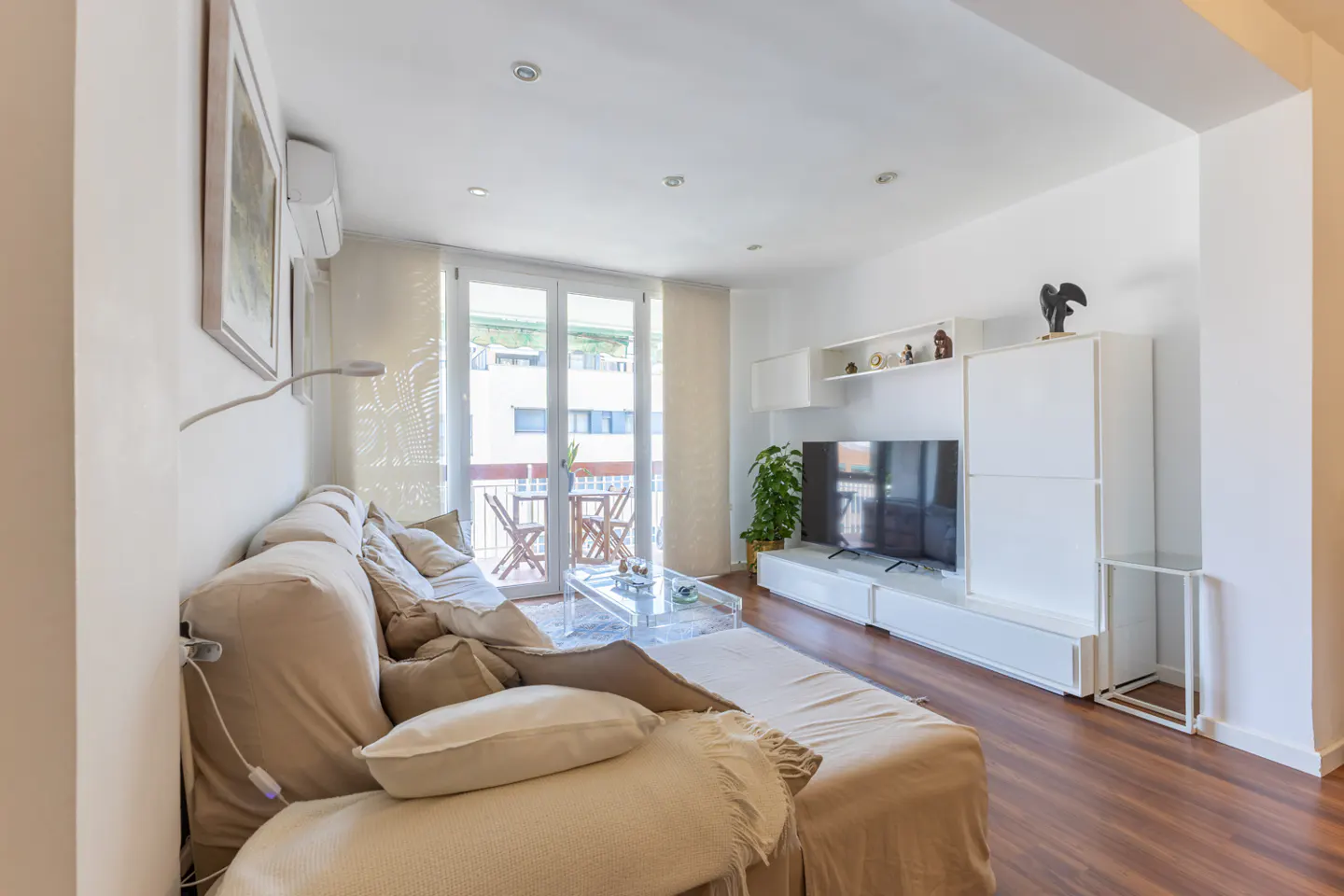 Bright living room with a beige sofa, glass table, and white entertainment center with a TV. Balcony with chairs visible.