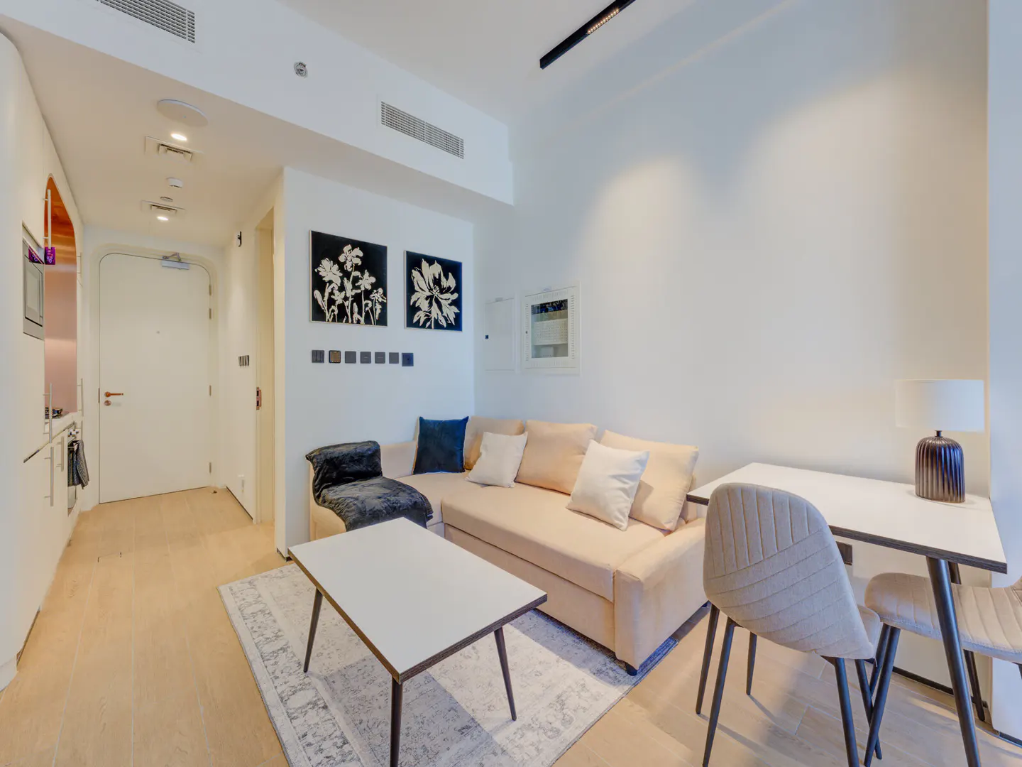 A bright studio apartment with a beige sofa, white coffee table, and a small desk with two chairs. Black and white floral art hangs above the sofa.
