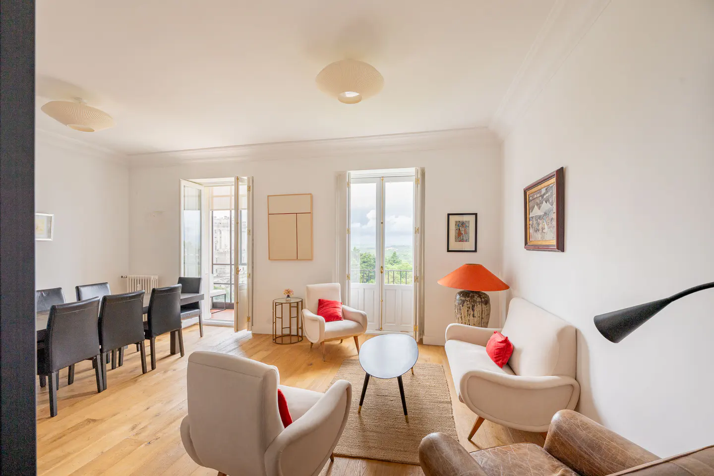 Bright living room with wood floors, white walls, and neutral furniture accented with red pillows. Balcony doors offer a view of greenery.