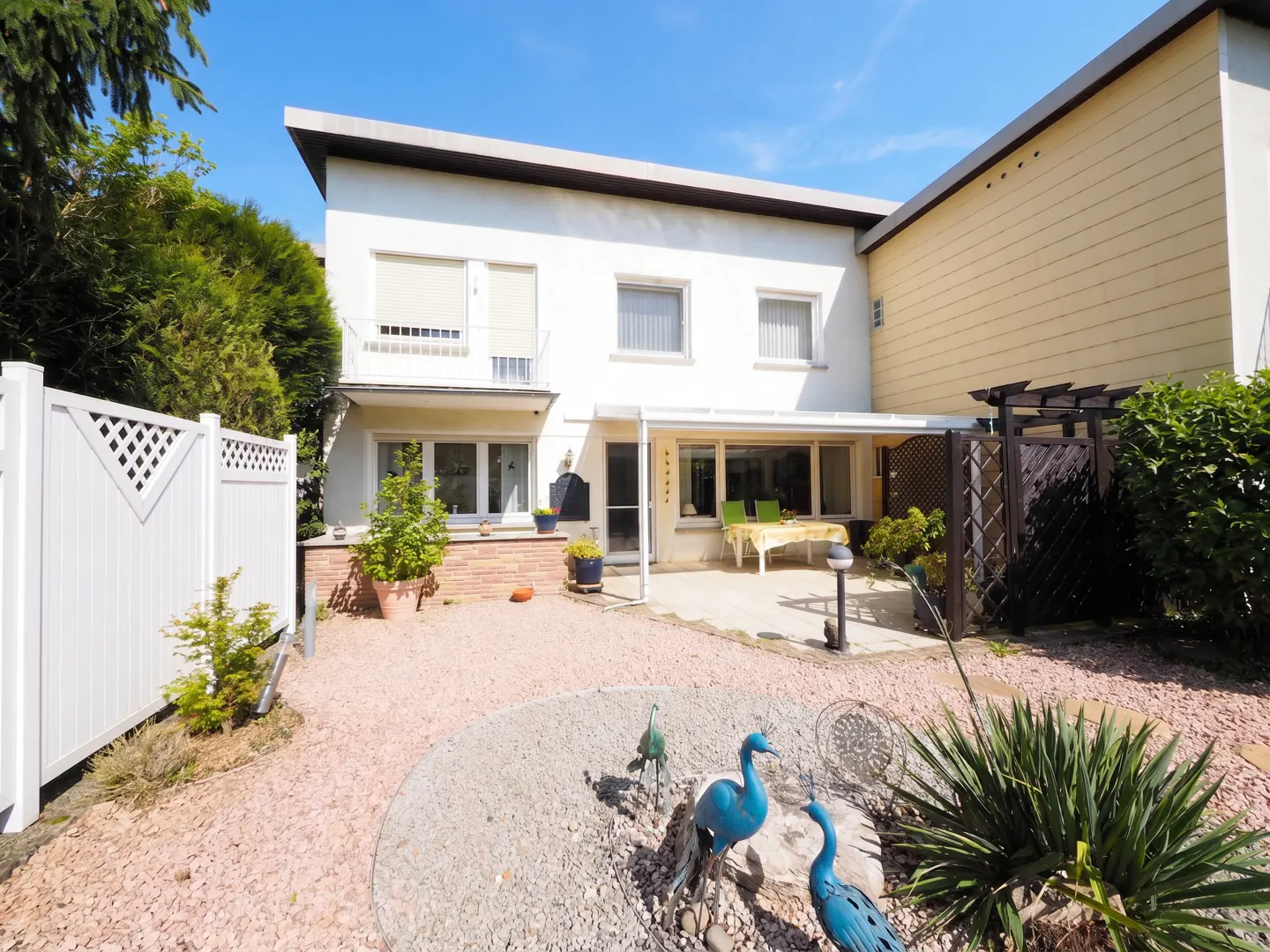 Backyard view of a two-story white house with a patio, gravel landscaping, and blue peacock statues. A white fence is on the left.