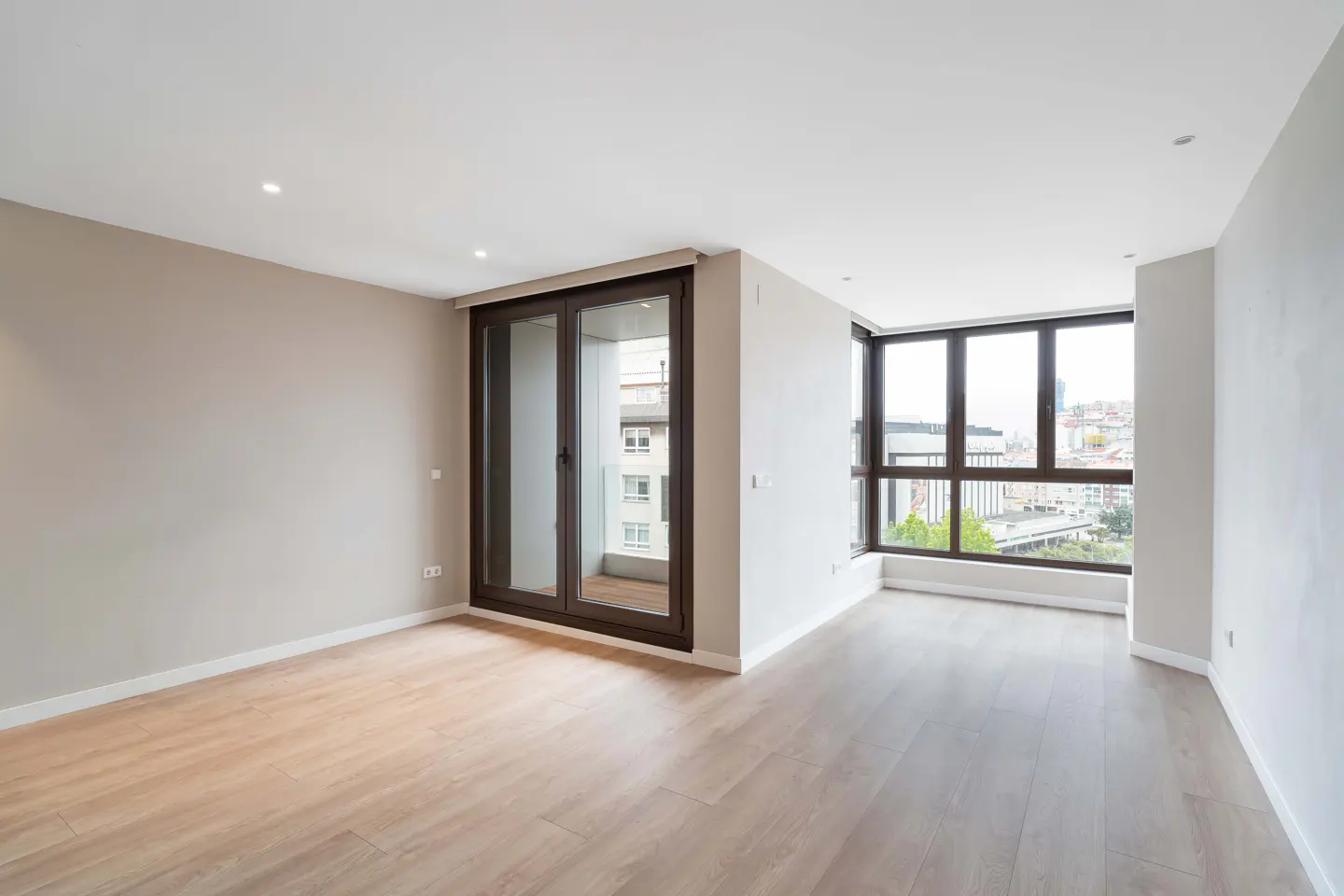 Bright, empty room with light wood floors, beige walls, and a large window with a city view. A sliding glass door leads to a small balcony.