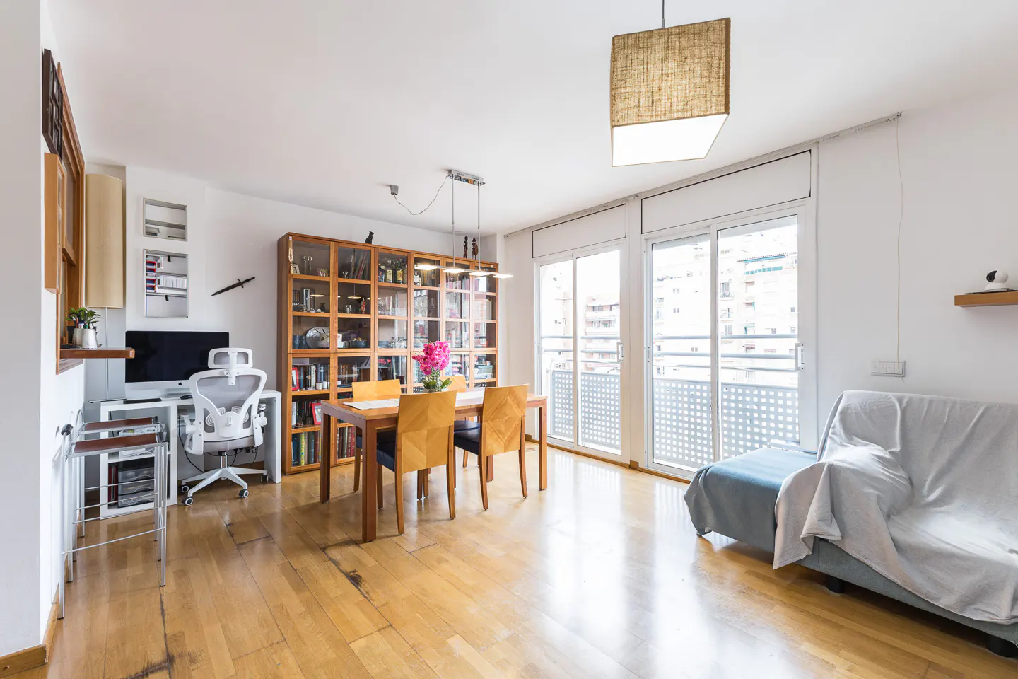 Bright, open-concept living space with wood floors, dining table, bookcase, desk, and sofa. Large windows offer city views.
