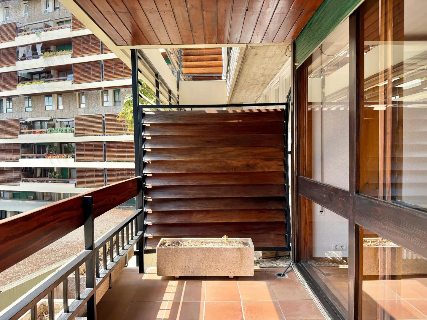 Balcony view with brown wood ceiling, railing, and privacy screen. A concrete planter sits on the orange tile floor. Building in background.