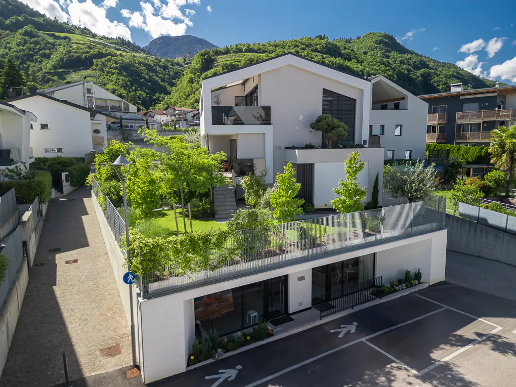 Modern white house with a green roof and parking lot, set against a backdrop of lush green hills and a blue sky.