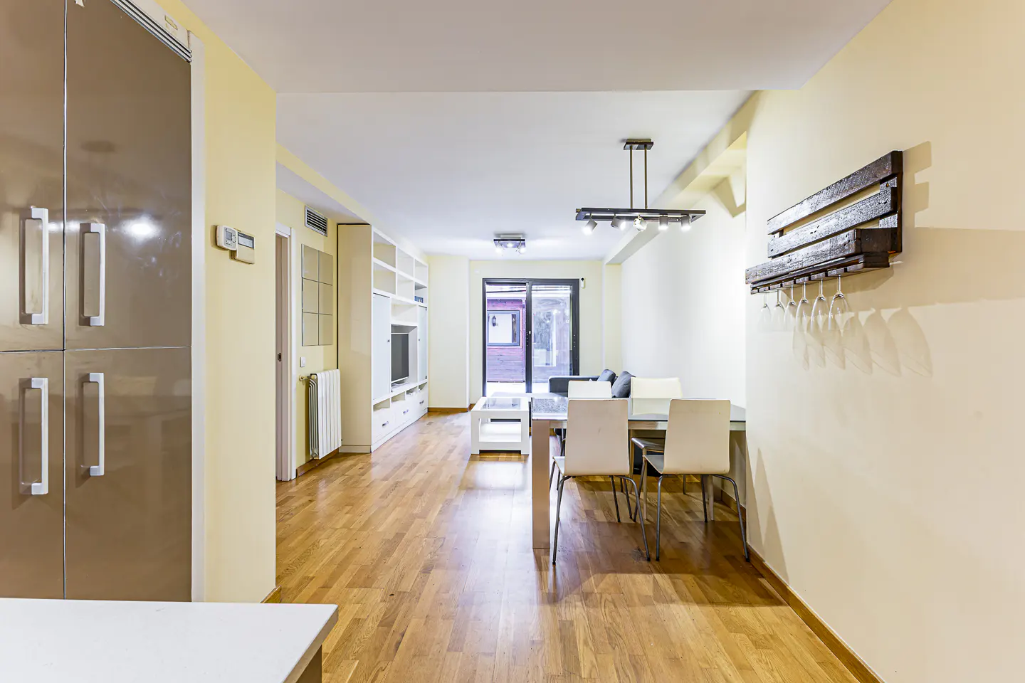 A bright, modern living room with hardwood floors, a dining table, and a large stainless steel refrigerator.