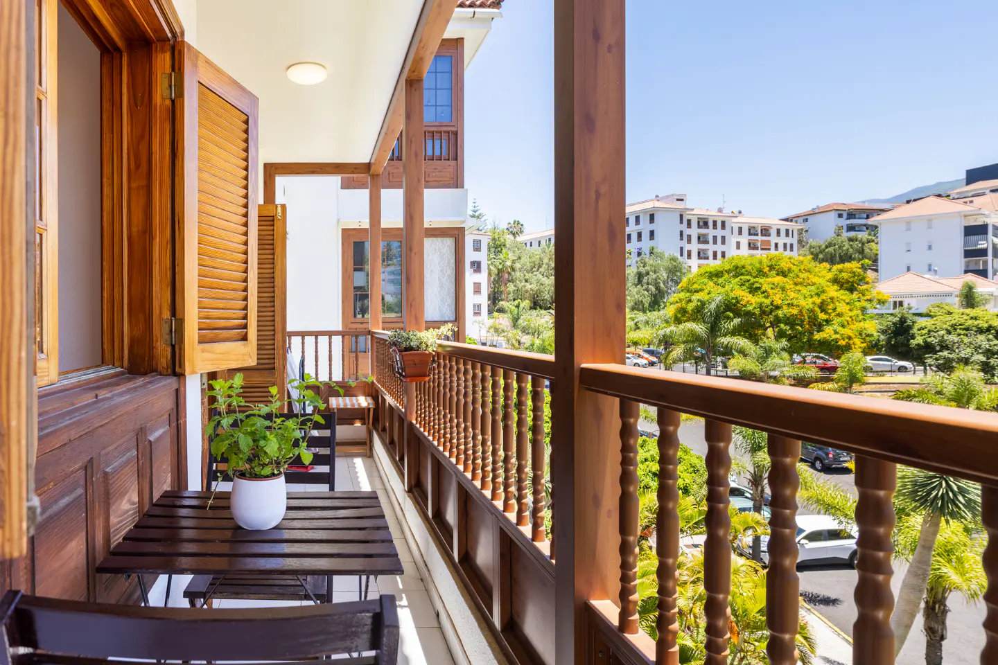 Wooden balcony with a small table, potted plant, and chairs, overlooking a sunny street with trees and white buildings.