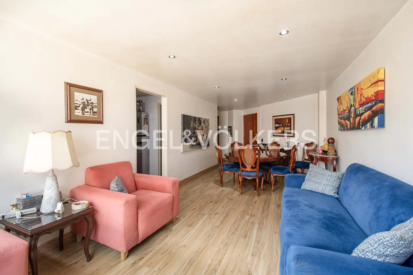 A bright living and dining area with a blue sofa, pink armchair, and round wooden dining table. Artwork adorns the white walls.