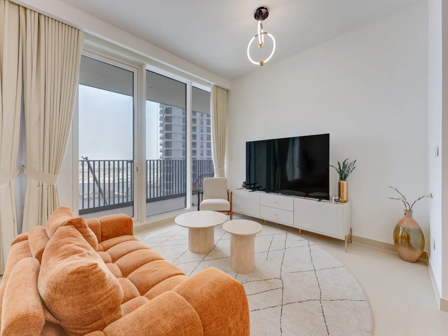 Bright living room with orange sofa, white rug, and TV. Balcony doors show city view. Modern light fixture overhead.