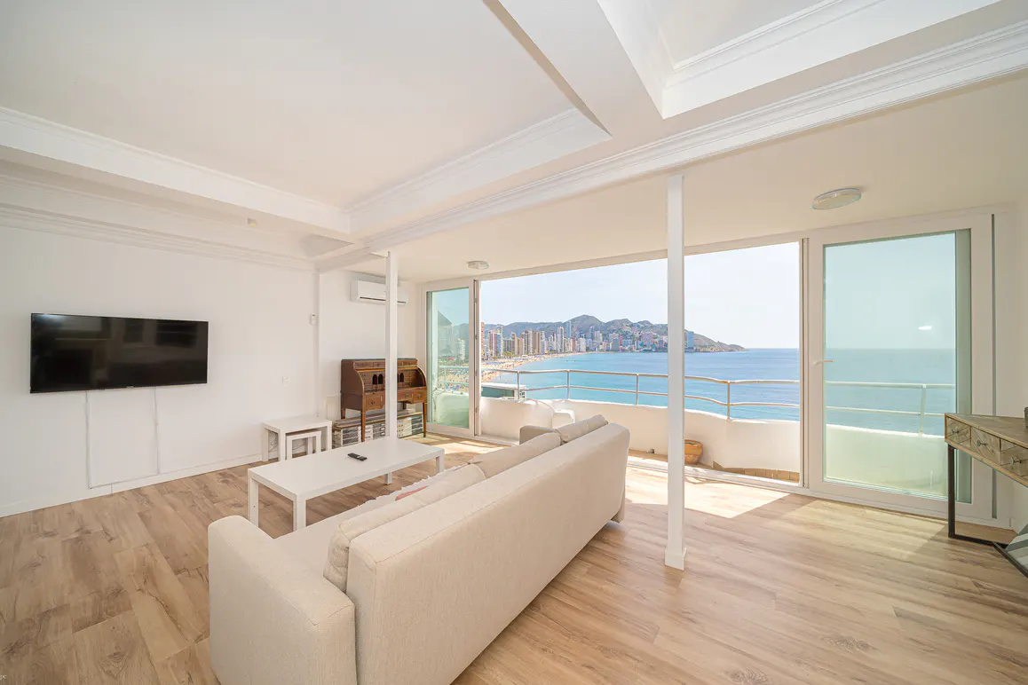 Bright living room with wood floors, white walls, and a large window overlooking a beach and ocean view. A white sofa and table are in the foreground.