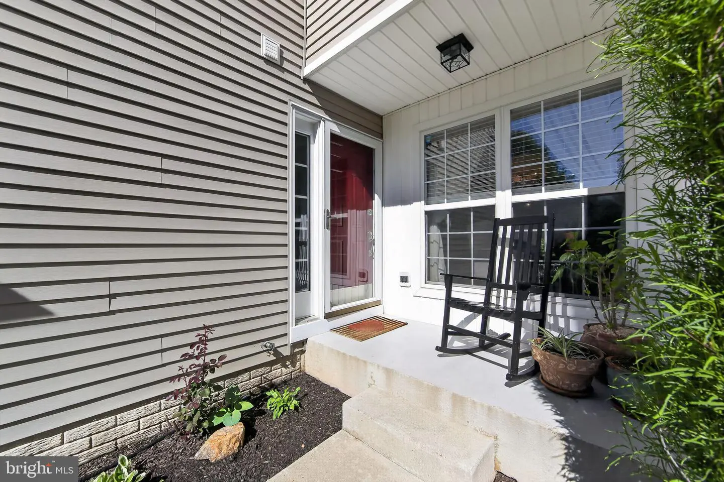 Home exterior with gray siding, red front door, and a black rocking chair on the porch. Windows with white trim and green plants.