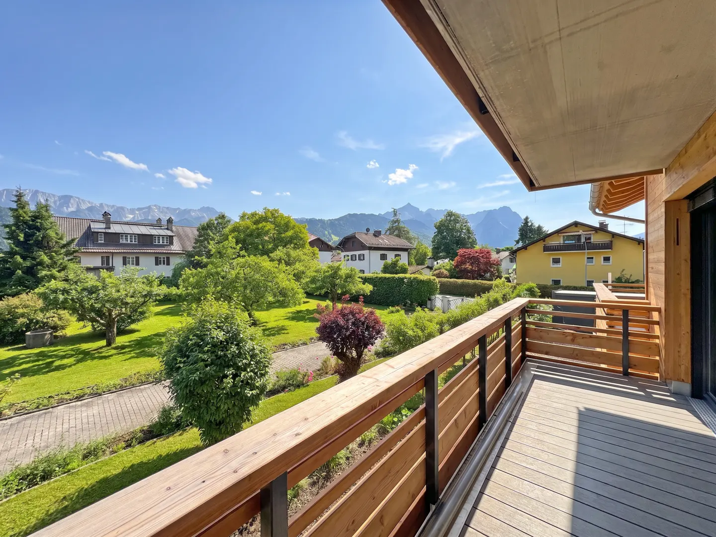 View from a wood balcony with a wood railing overlooking green lawns, houses, and mountains under a blue sky.