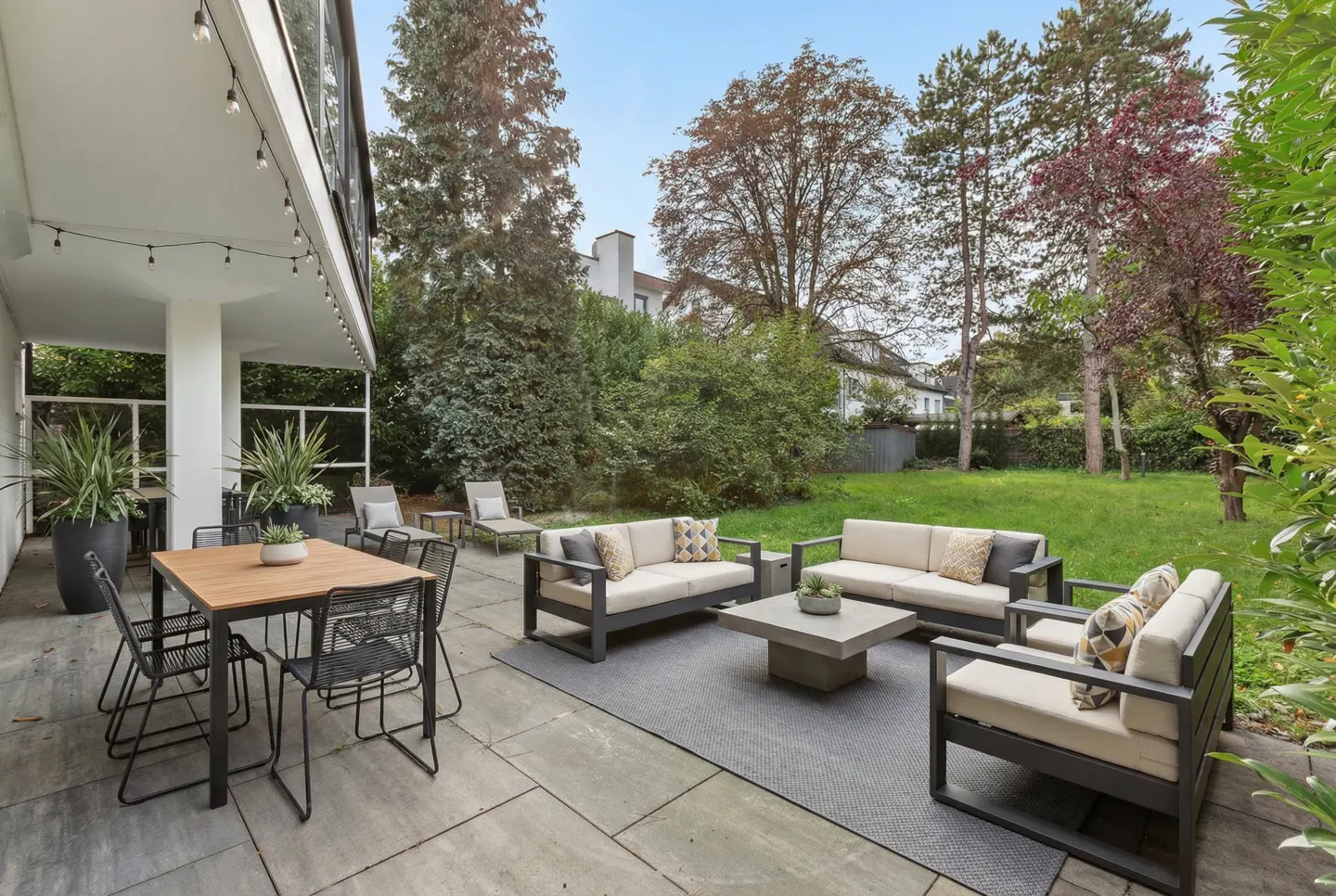 Outdoor patio with dining table, sofas, and chairs on a stone patio. Green lawn and trees in the background.