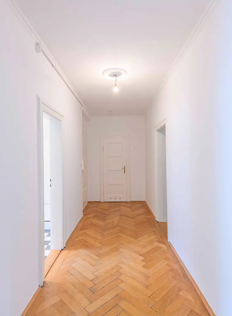 Bright hallway with herringbone wood floors, white walls, and doors. A light fixture hangs from the ceiling.