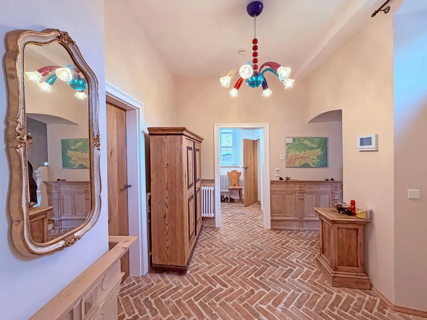 Hallway with brick floors, wooden furniture, and a colorful chandelier. A large ornate mirror hangs on the wall.