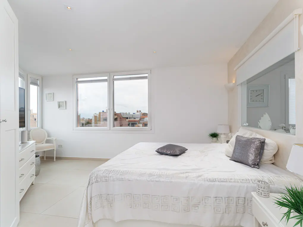 Bright, all-white bedroom with a large bed, gray pillows, and a view of the city through the windows. A white wardrobe and chair are on the left.