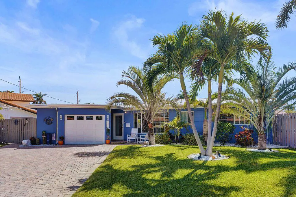 Blue single-story house with a white garage door, palm trees, and a brick driveway under a blue sky.