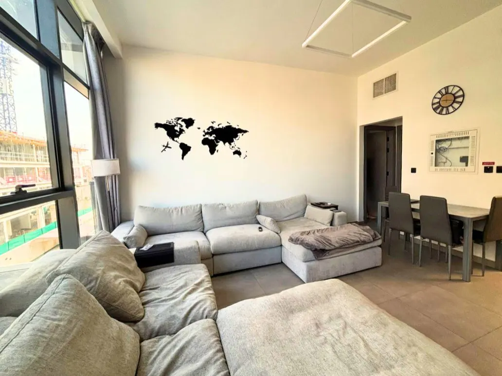 Living room with a large gray sectional sofa, dining table, and a black world map wall decal. Natural light from a large window.