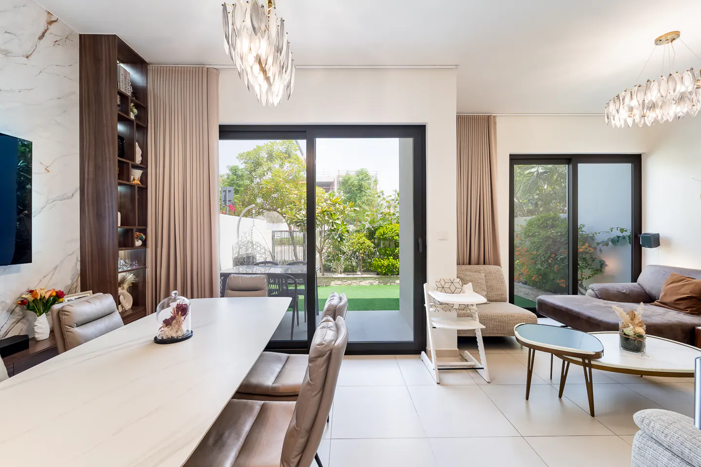 Bright, modern living and dining area with a long white table, brown chairs, and sliding glass doors to a green backyard. Chandeliers hang above.
