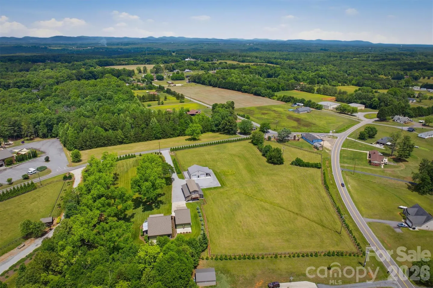 Aerial view of a property with a house, green lawn, trees, and a road. Mountains are visible in the distance under a blue sky.