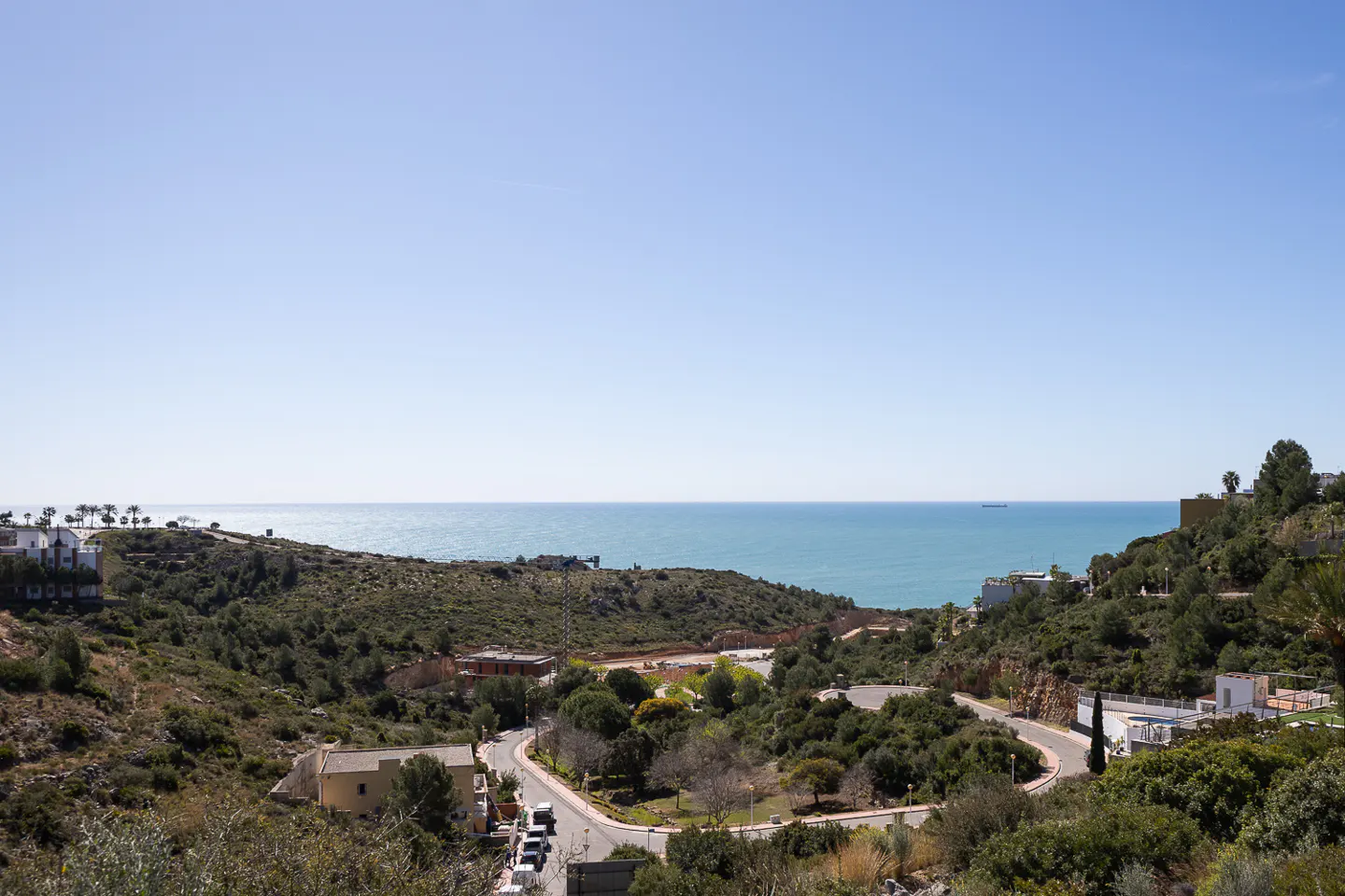 Scenic view of a winding road through green hills, leading to a blue ocean under a clear sky. Houses are scattered among the trees.