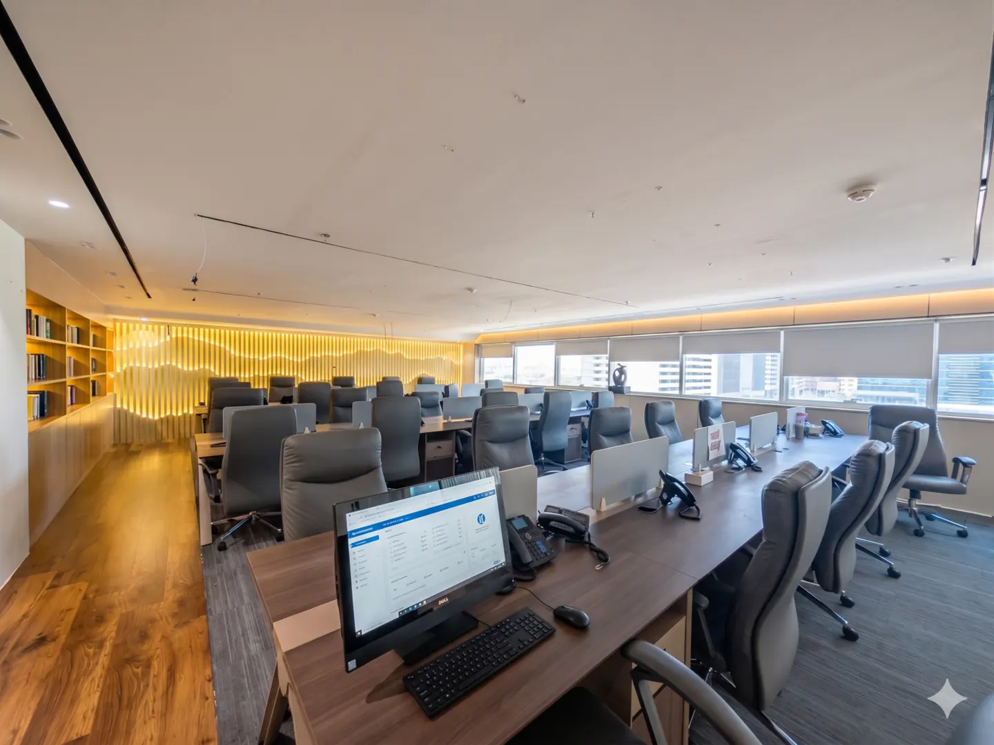Bright office space with rows of desks, computers, and gray chairs. A bookshelf and decorative wall are on the left. Windows show a city view.