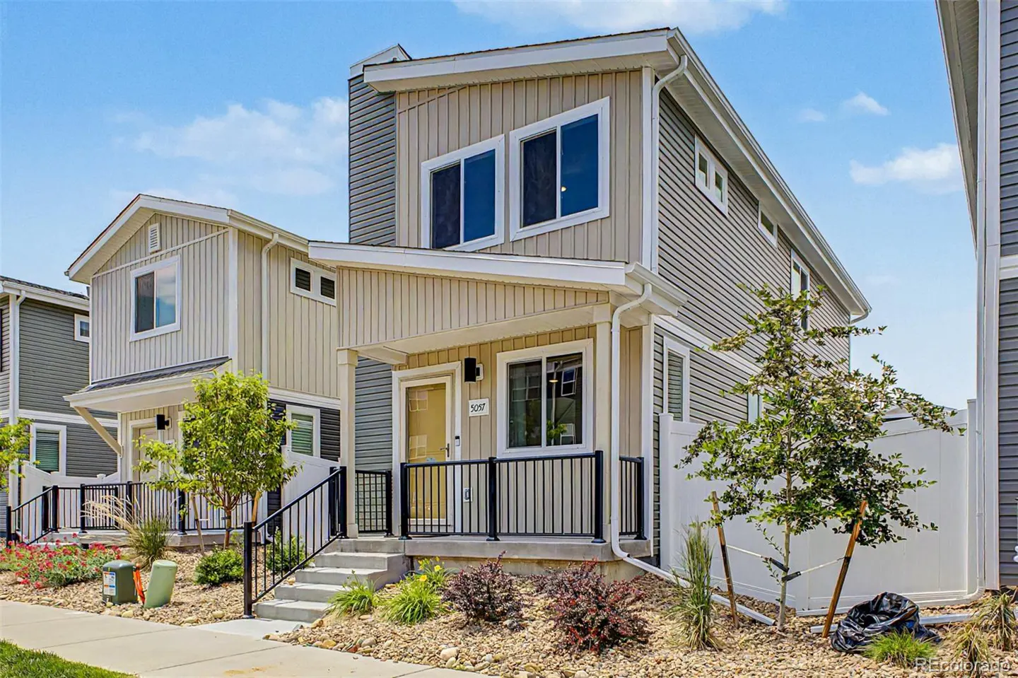 Two-story townhouse with beige and gray siding, black railings, and a small front yard with rocks and plants. Blue sky in the background.