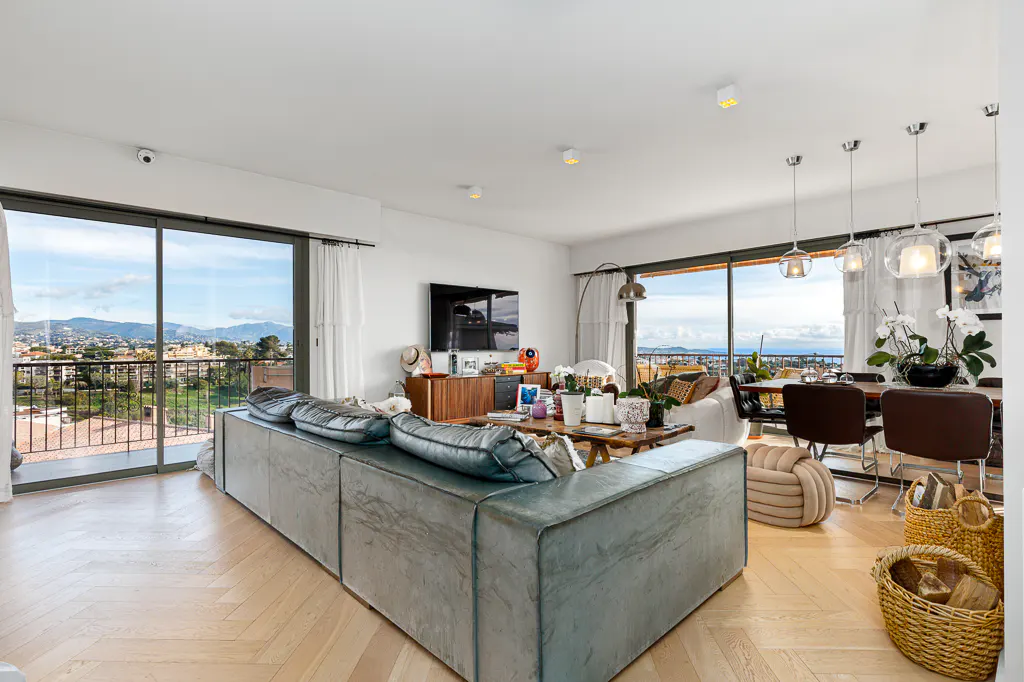 Bright, modern living room with a large gray couch, wood floors, and floor-to-ceiling windows with a city view.