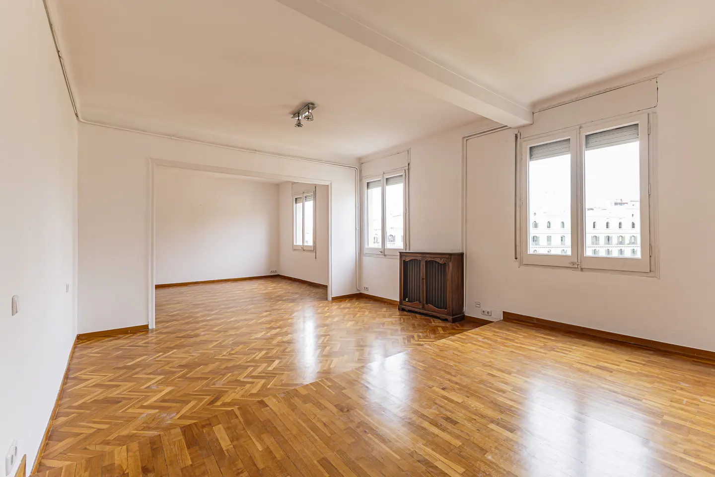 Bright, empty living room with herringbone wood floors, white walls, large windows, and a wooden radiator cover.
