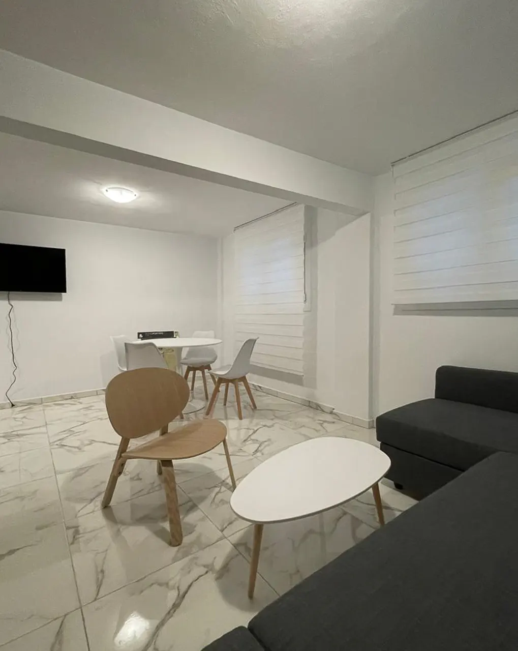 A modern living room with white walls, marble floors, and a gray sectional sofa. A wooden chair and white coffee table sit in the center. A dining table with chairs is in the background.
