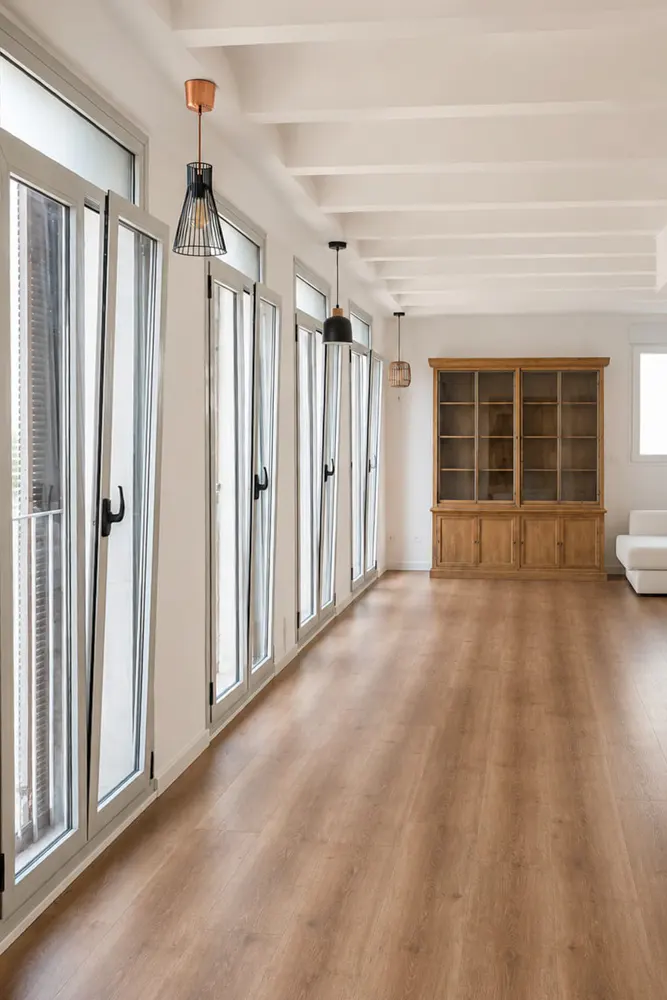 Bright, empty room with wood floors, white walls, and exposed ceiling beams. Multiple windows line the left wall, and a large wooden cabinet sits at the back.