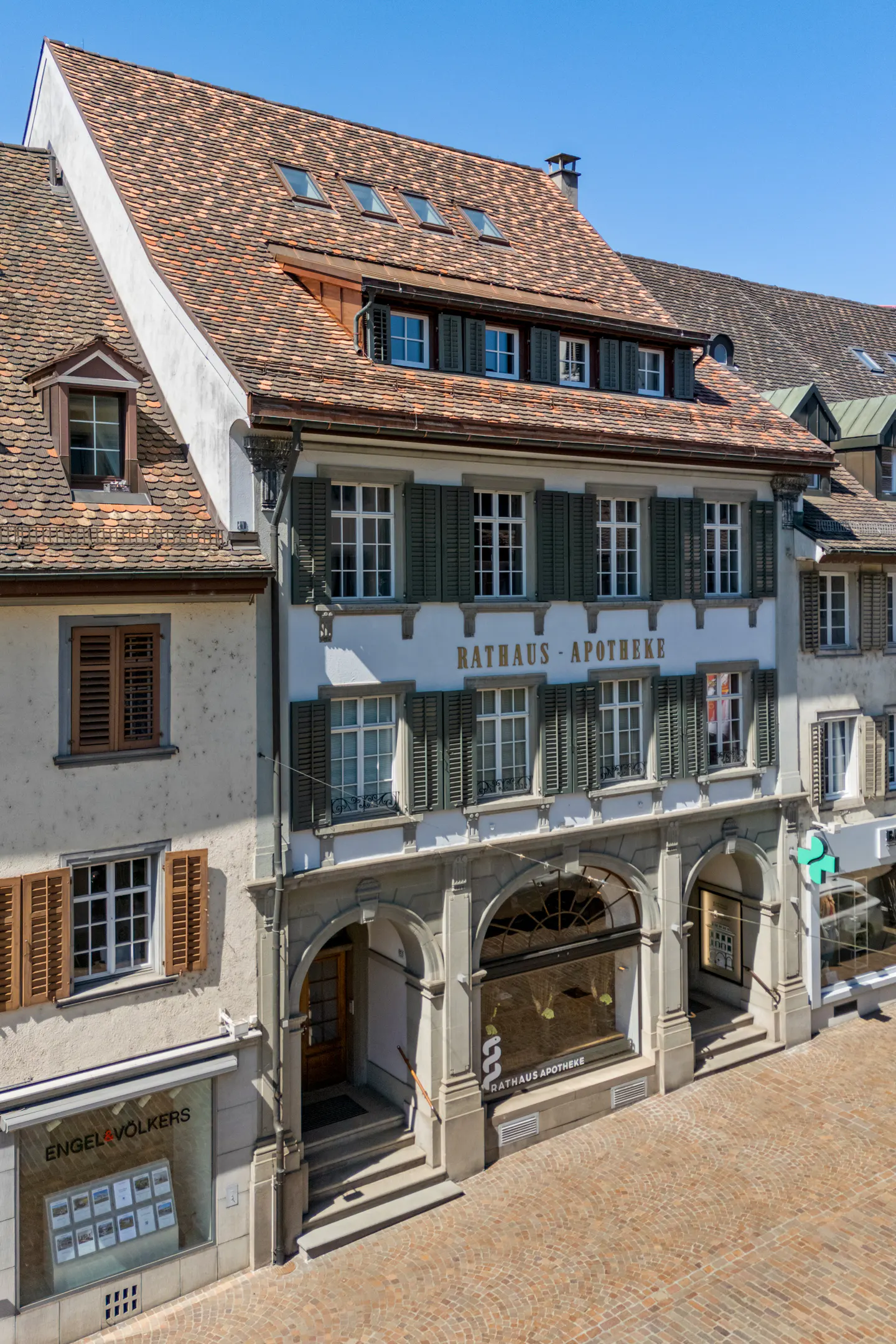 Exterior view of a three-story building with "RATHAUS APOTHEKE" sign, green shutters, arched windows, and a red-tiled roof.