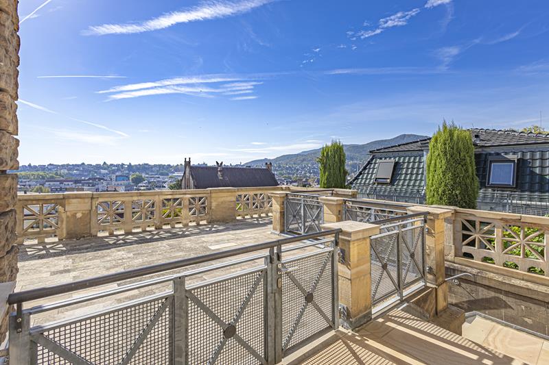 Spacious stone terrace with metal railings overlooking a city, distant mountains, and a bright blue sky. [AI-generated text]