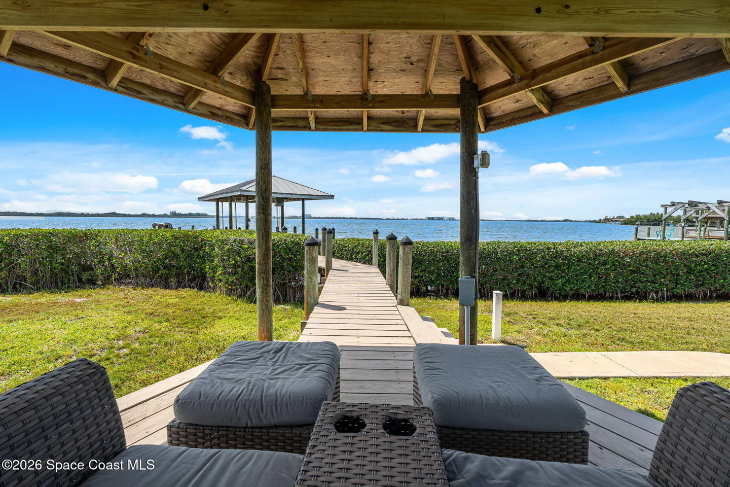 View from a gazebo with gray cushioned seating, looking out to a wooden dock, water, and a distant gazebo under a blue sky.