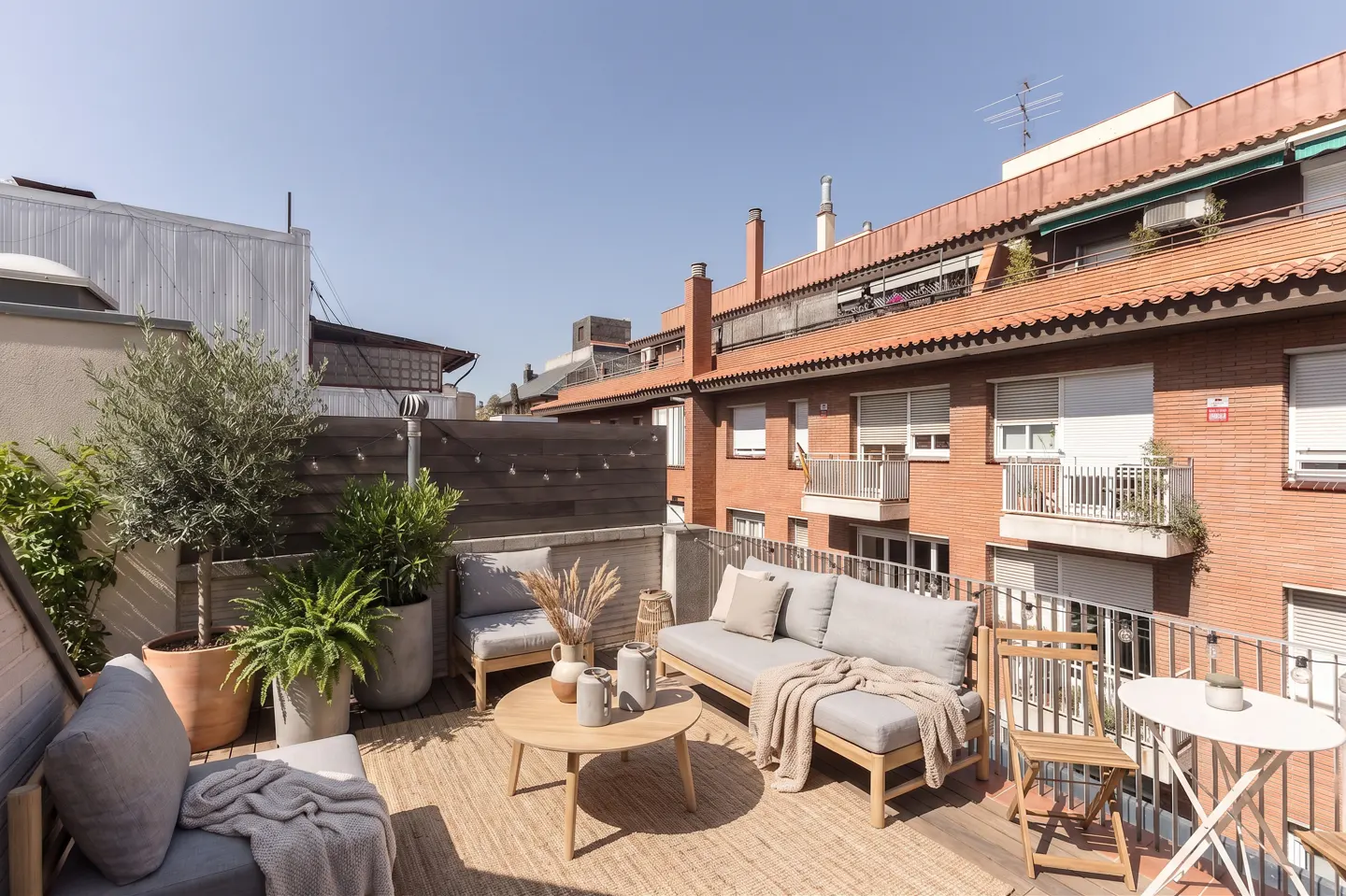 Outdoor patio with gray cushioned seating, a round wooden table, and potted plants on a jute rug, with brick buildings in the background.
