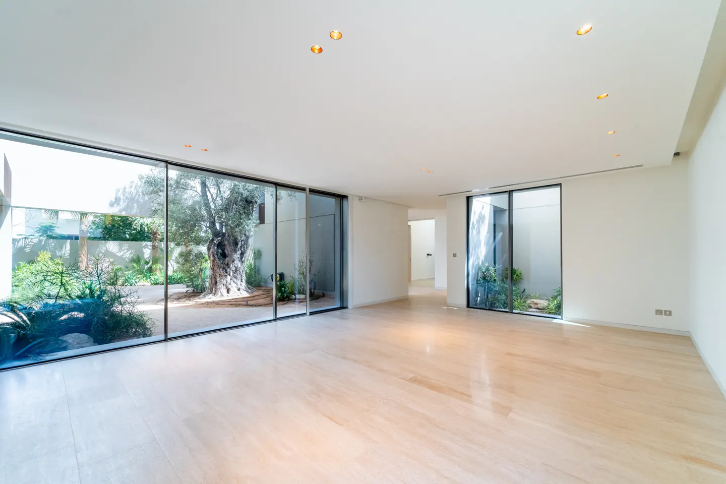 Bright, empty room with marble floors and white walls. Large glass doors reveal a courtyard with a tree and greenery.