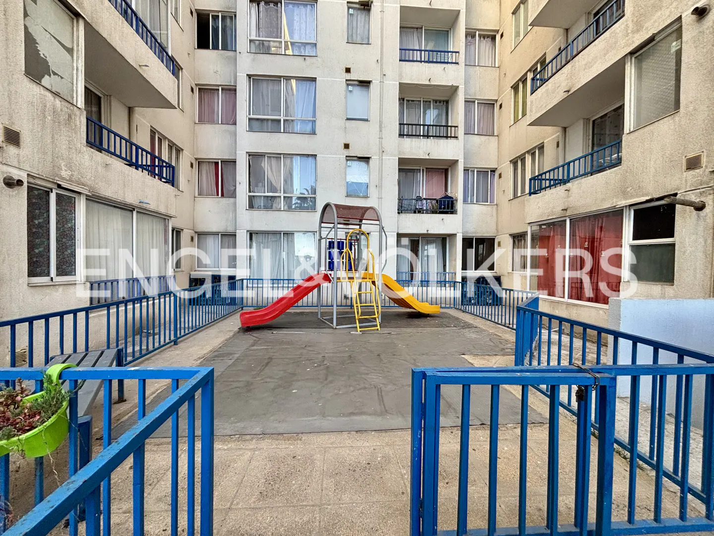 Apartment complex courtyard with a playground. Red and yellow slides sit on a gray surface, surrounded by blue railings. Balconies and windows above.