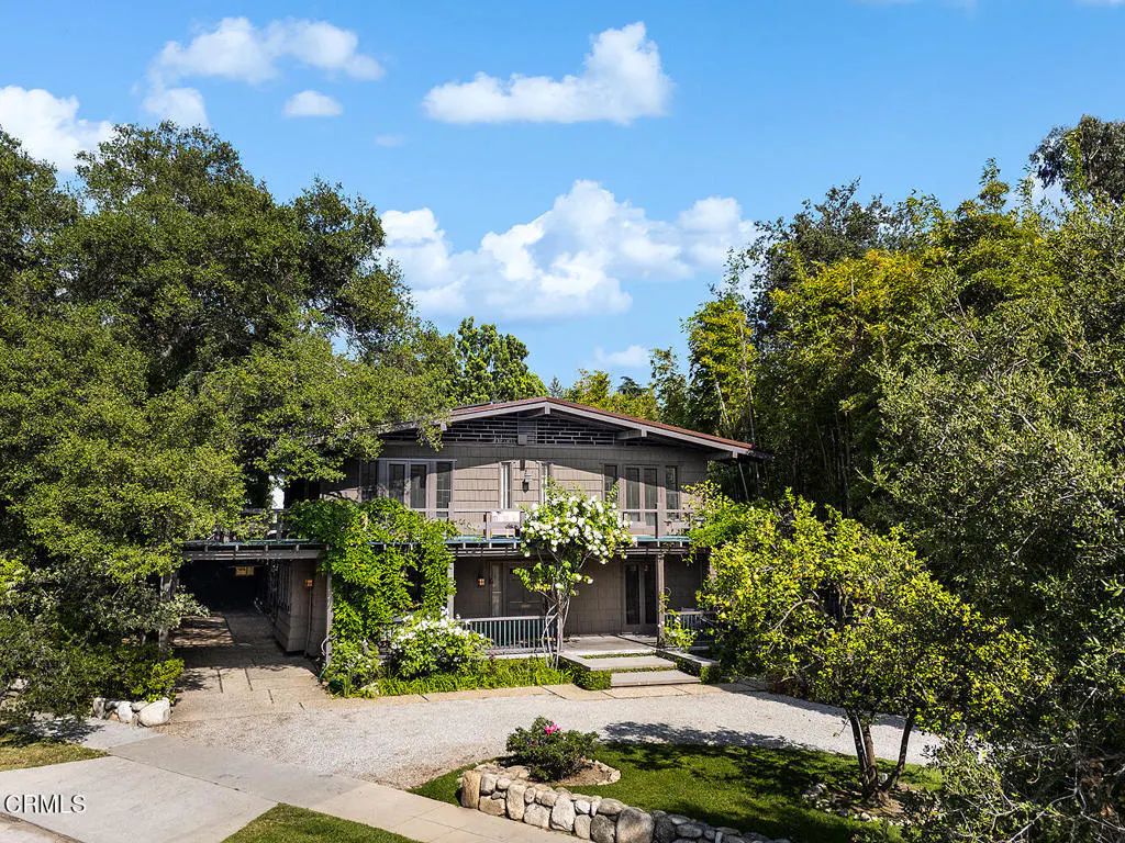 Exterior view of a two-story gray house with a brown roof, surrounded by lush green trees and a blue sky with white clouds.