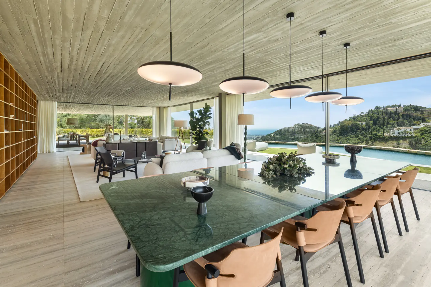 A modern dining room with a green marble table, tan chairs, and pendant lights. Large windows offer a view of the pool and landscape.