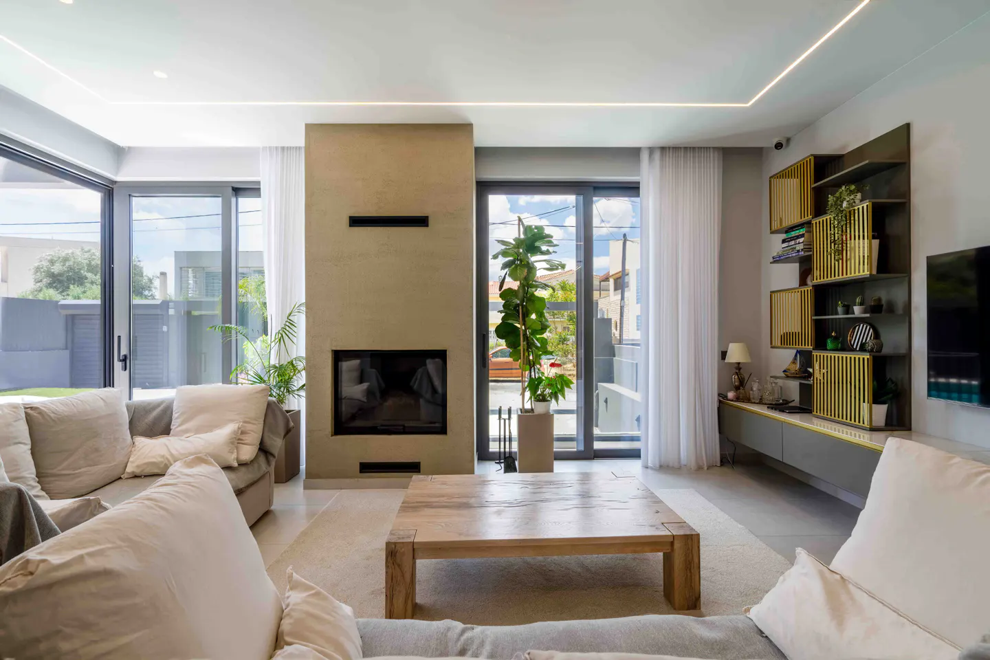 Living room with a beige fireplace, a wooden coffee table, and a gray sofa. Large windows let in natural light. A modern bookshelf is on the right.
