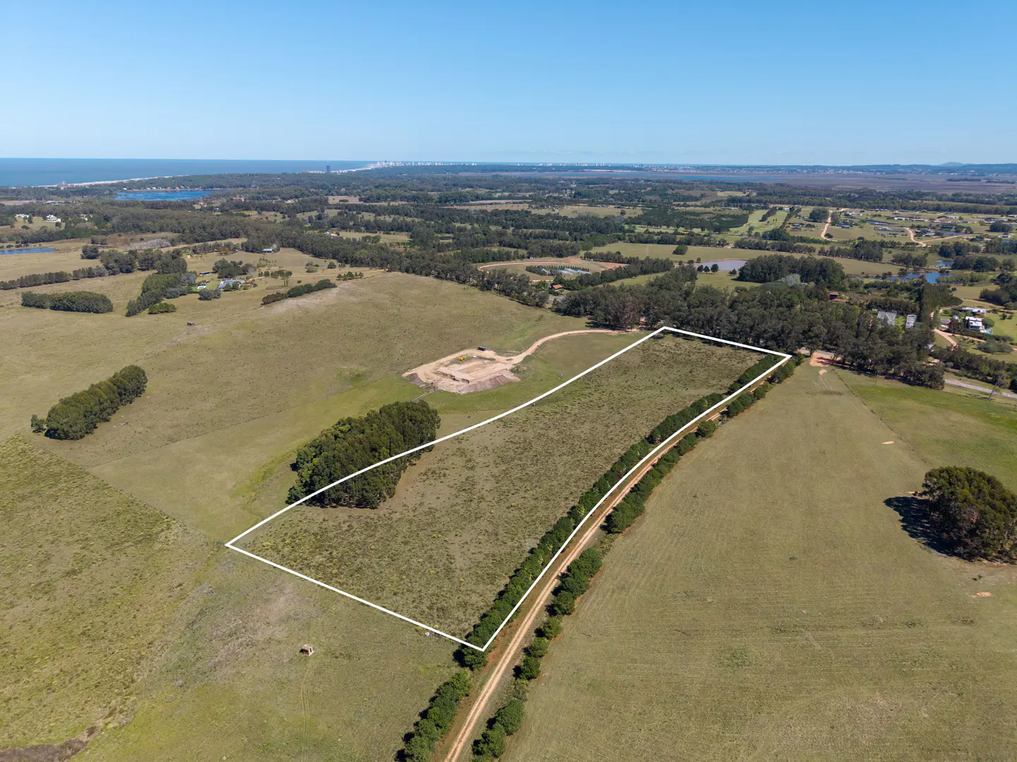 Aerial view of a large, rectangular plot of land outlined in white, surrounded by green fields and trees under a clear blue sky.