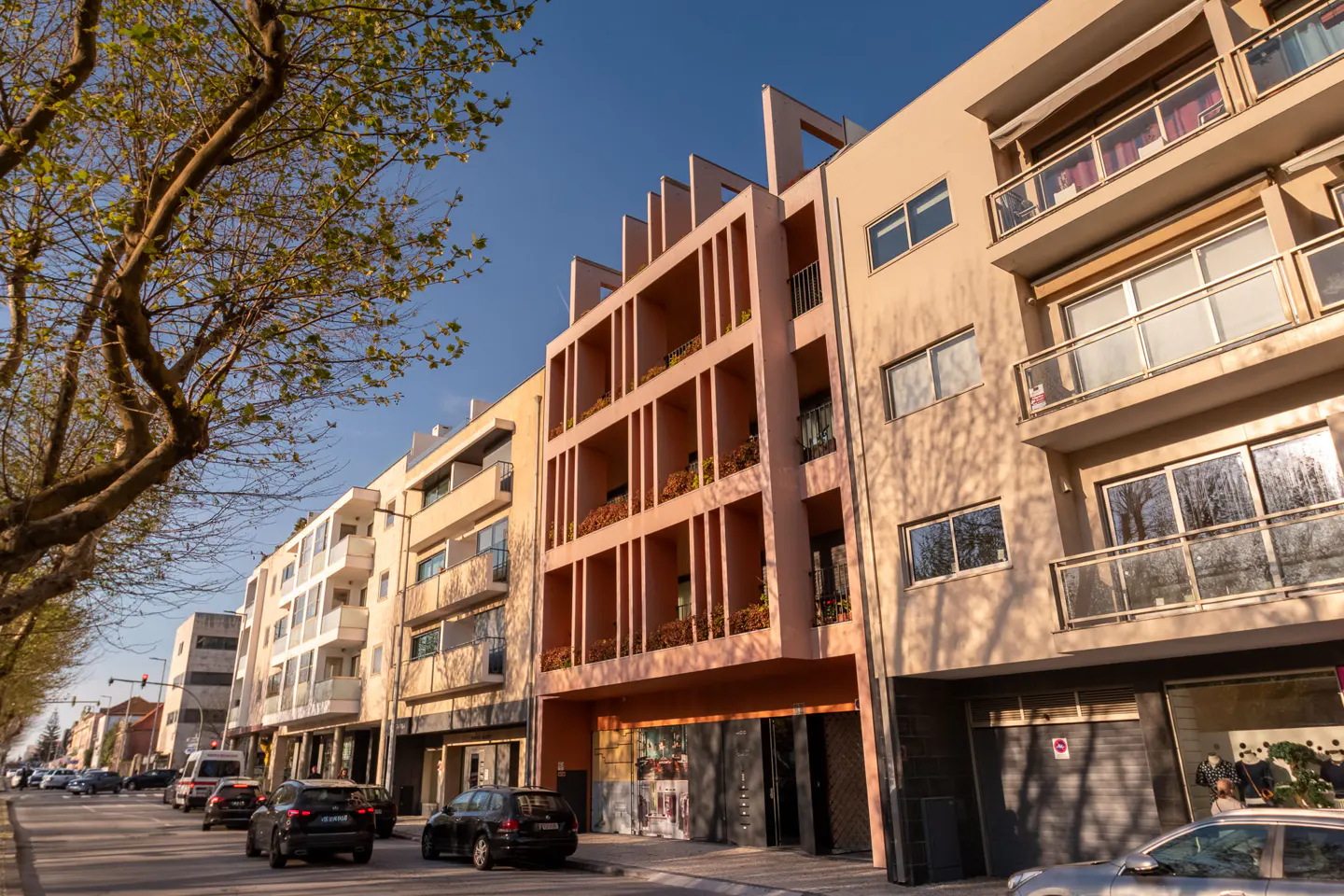 Street view of modern, multi-story apartment buildings in shades of beige and pink, with cars on the road.