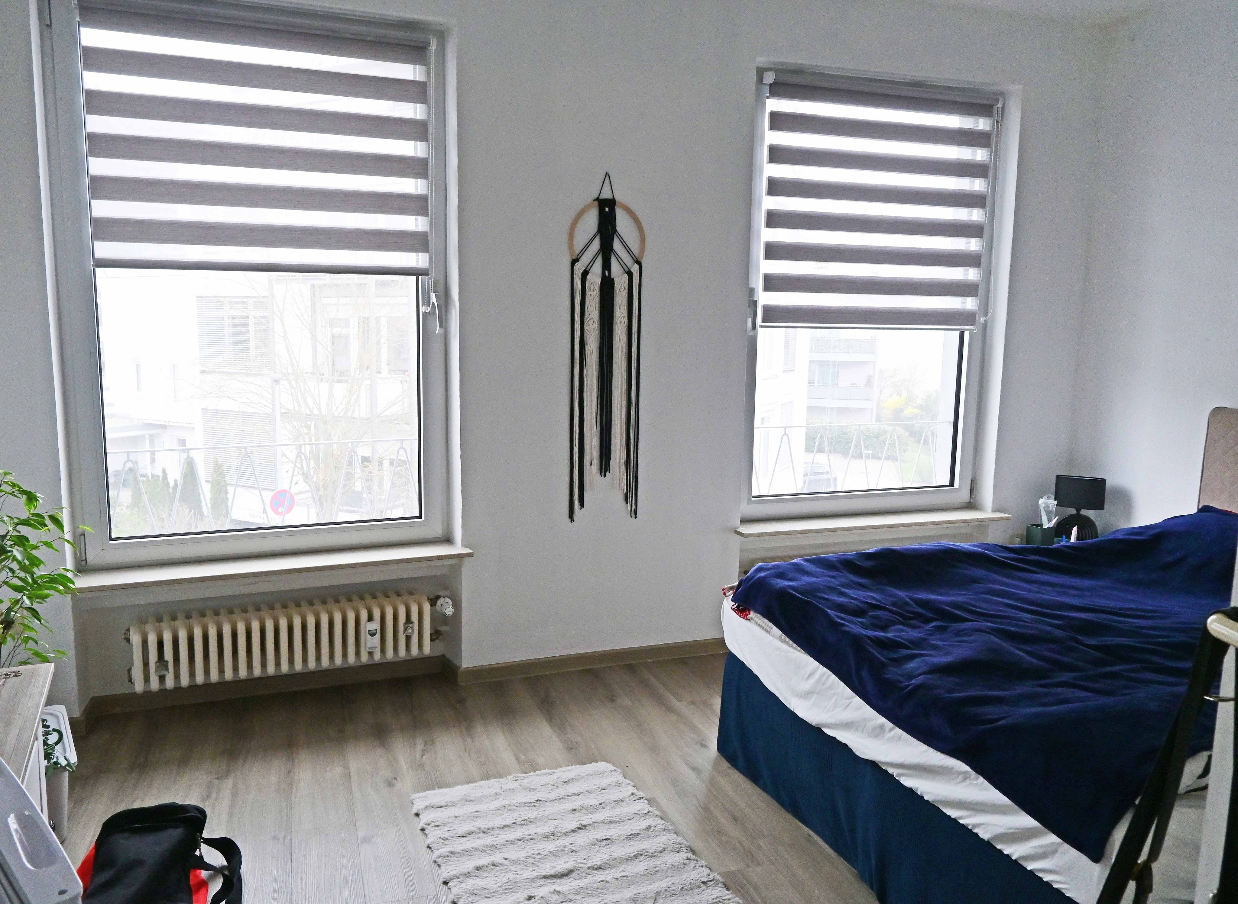 Bedroom with two windows covered by gray blinds, a bed with blue bedding, and a macrame wall hanging. [AI-generated text]