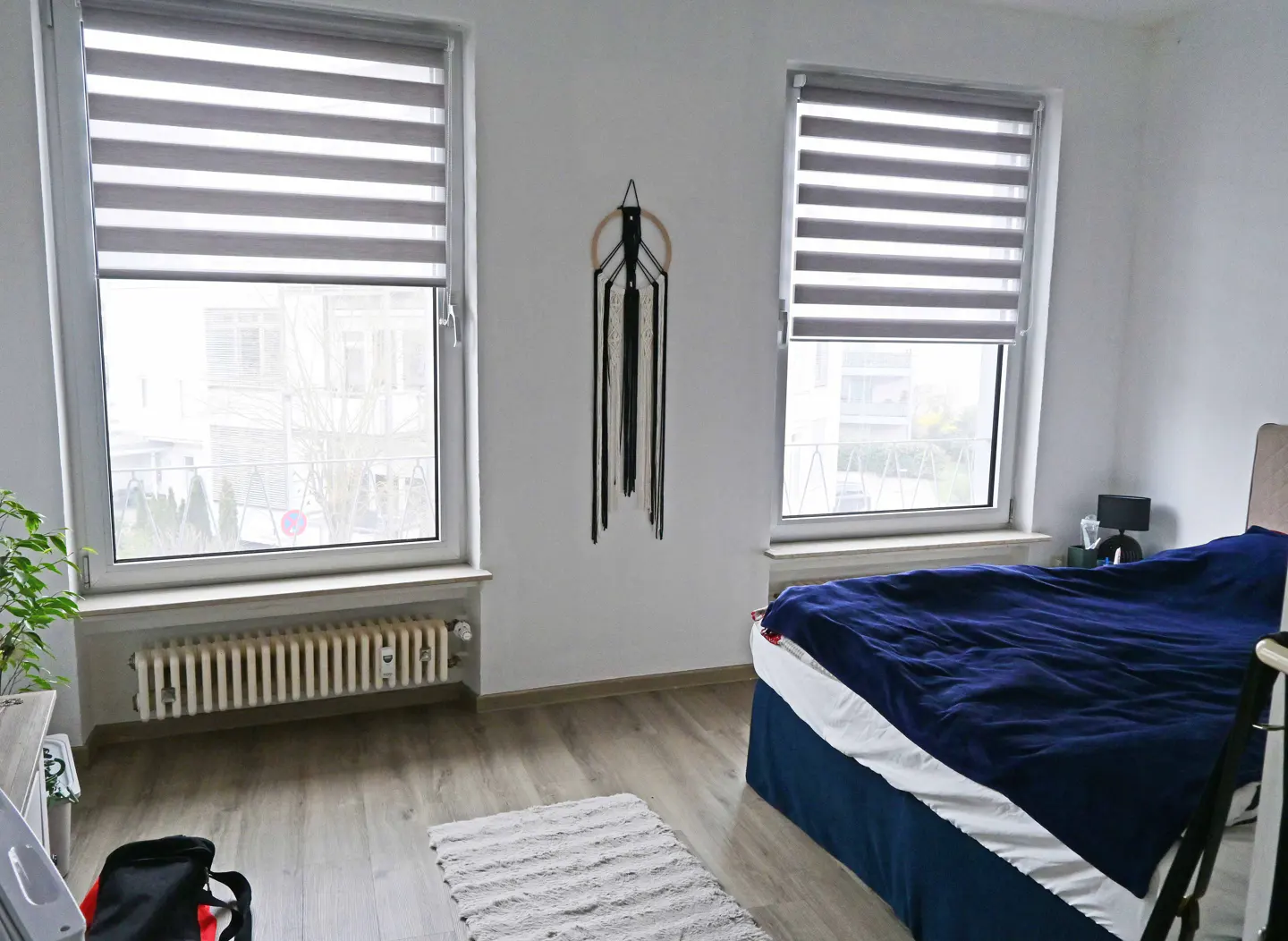 Bedroom with two windows covered by gray blinds, a bed with blue bedding, and a macrame wall hanging.