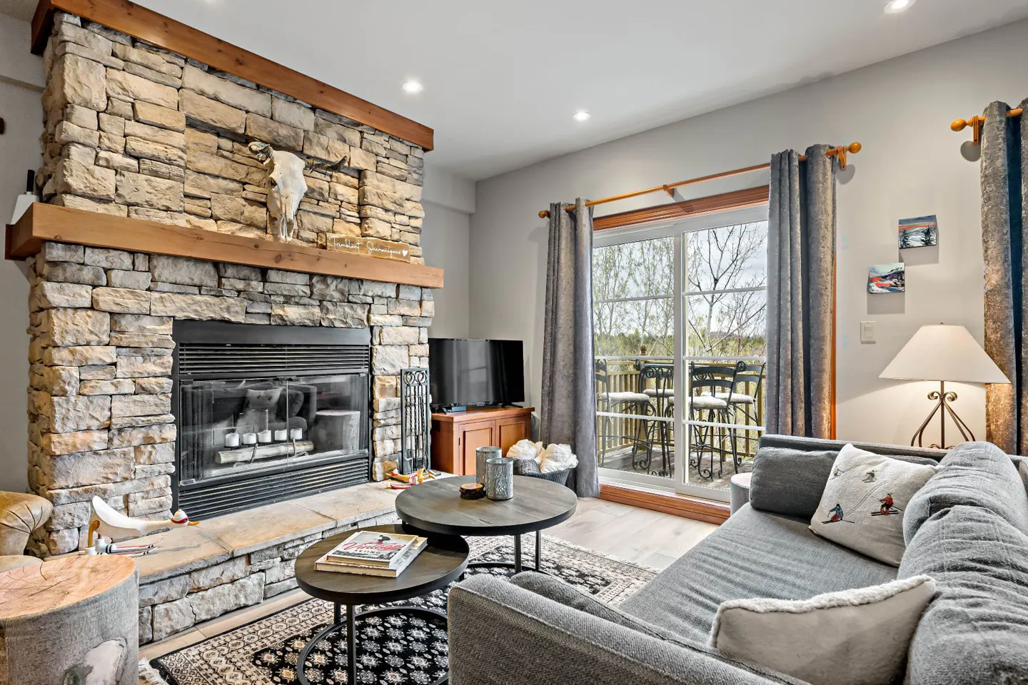 Living room with stone fireplace, wooden mantle, and skull. Gray sofa, round tables, and sliding glass doors to a deck.