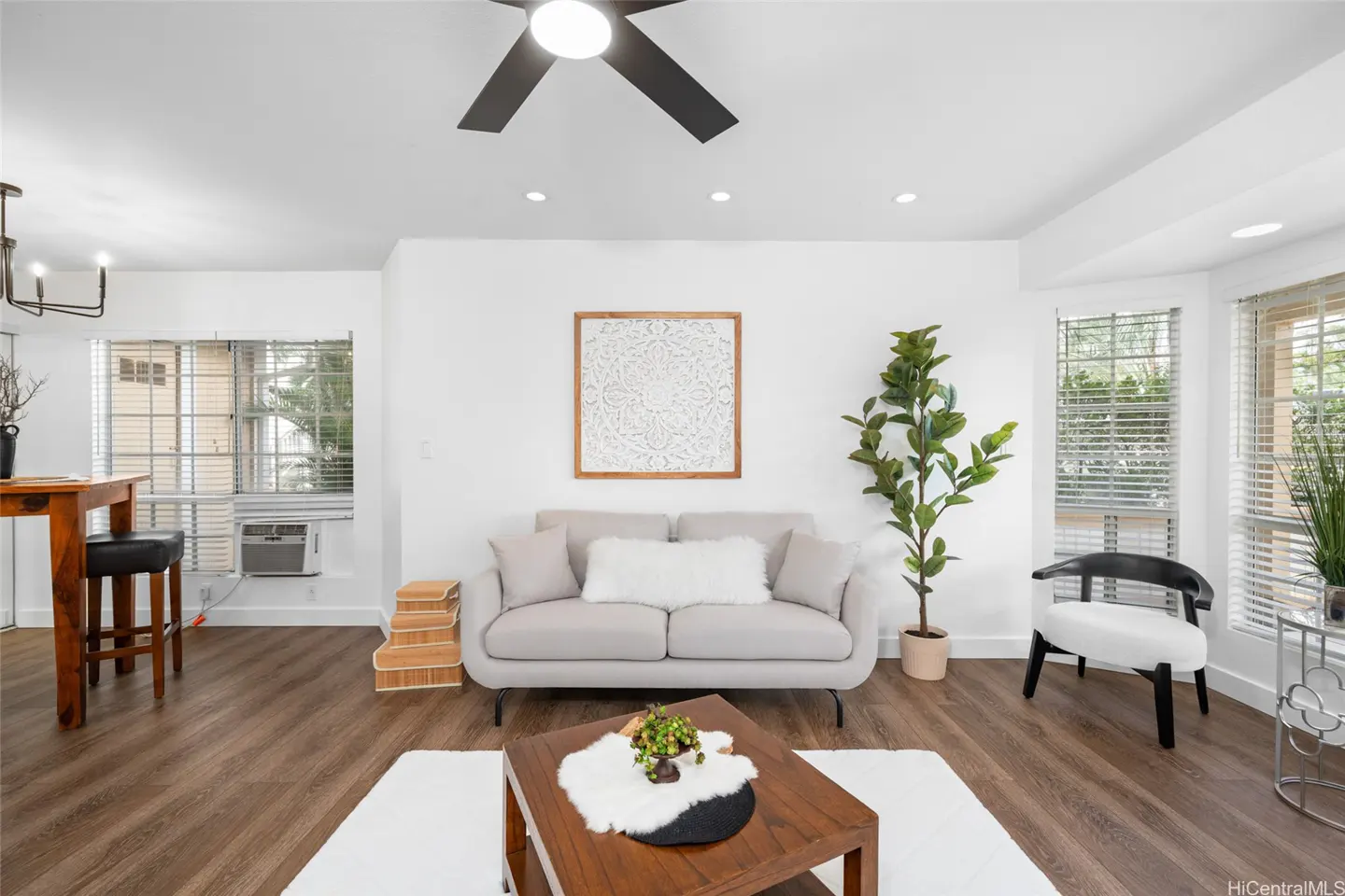 Bright living room with white walls, wood floors, and a gray sofa. A ceiling fan and windows provide light. A wooden coffee table sits on a white rug.