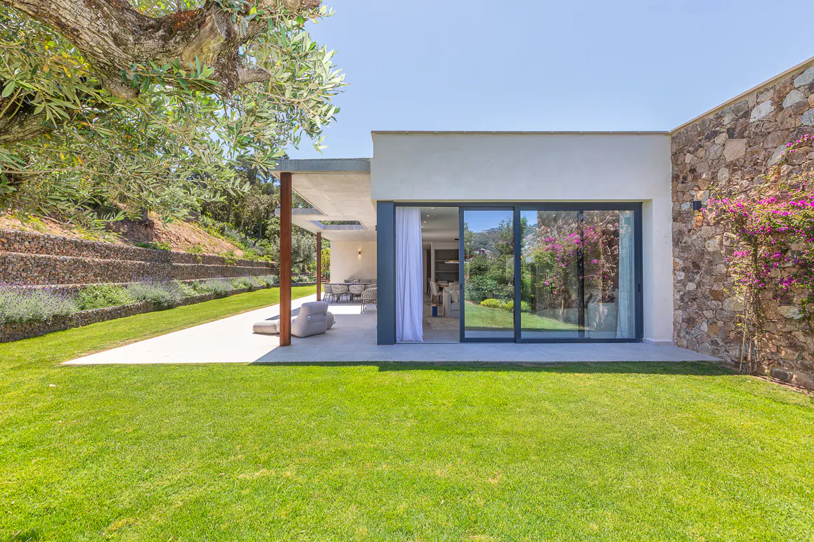 Modern home with stone wall, glass sliding doors, and patio overlooking a green lawn under a clear blue sky.