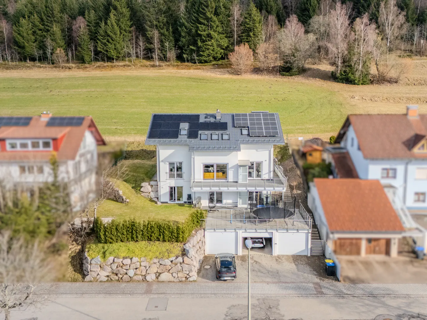 Aerial view of a white multi-story house with solar panels on the roof, a terrace, and a garage, set against a forest.