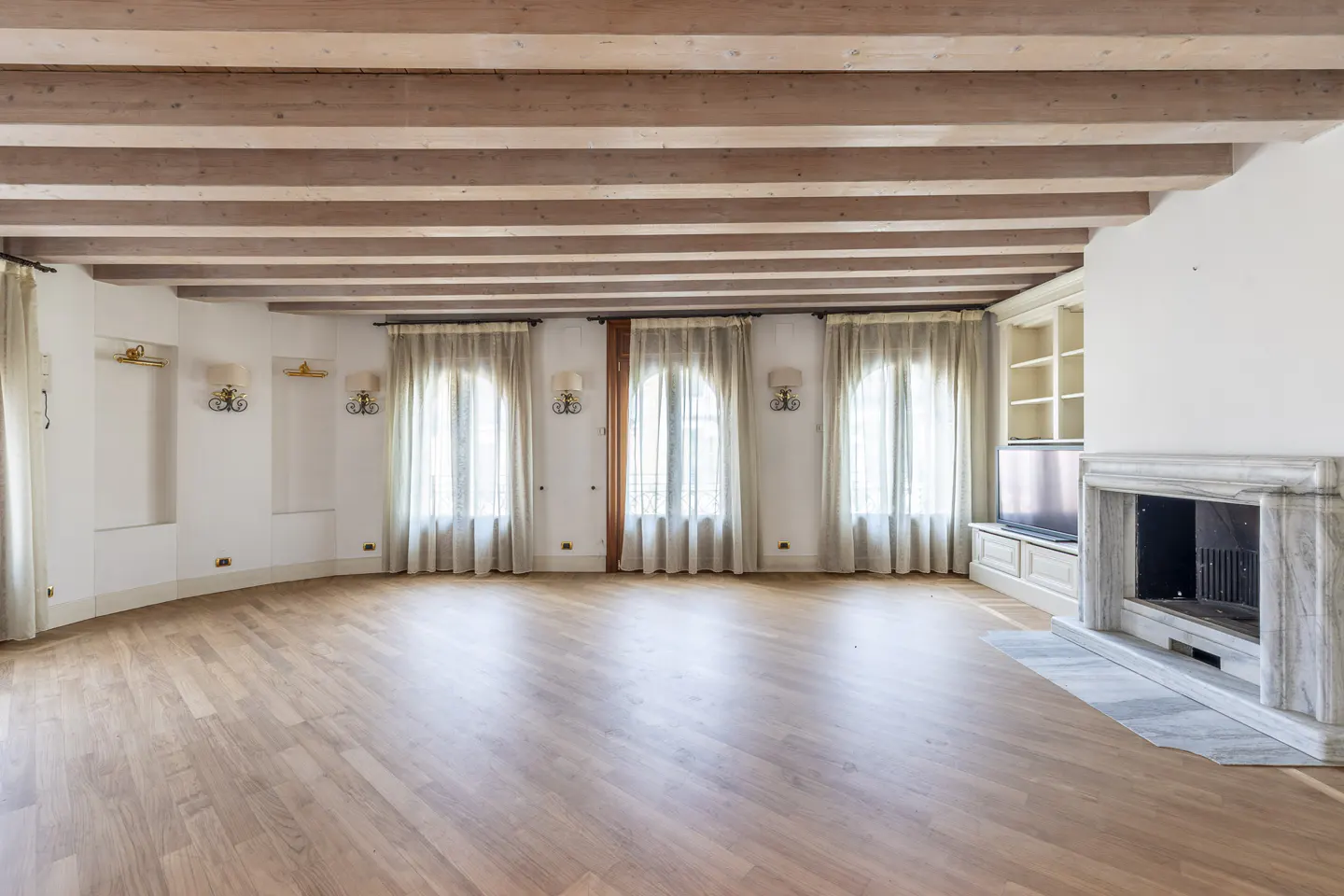 Bright, empty living room with wood floors, beamed ceiling, fireplace, and three arched windows with sheer curtains.