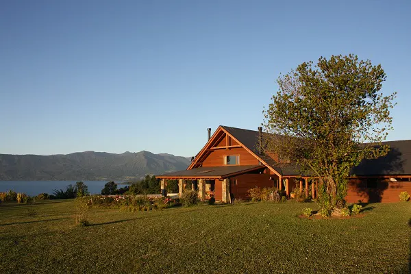 A brown wooden house with a porch sits on a green lawn, overlooking a lake and mountains under a blue sky.